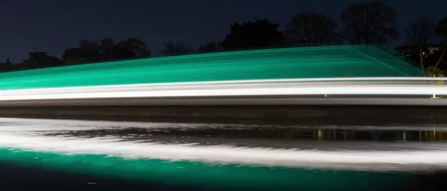 Long-exposure emerald and white light trails stretch across a night scene, mirrored on still water below