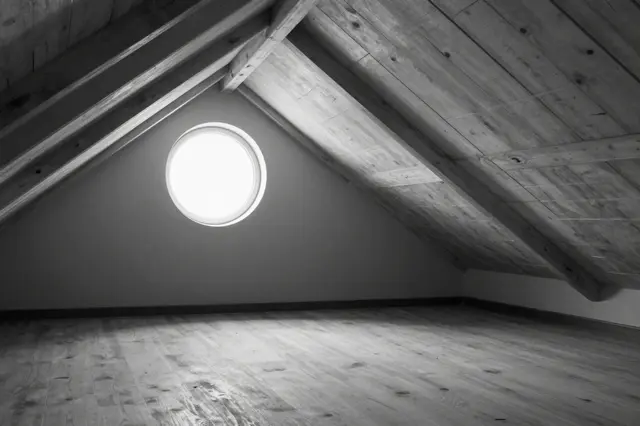 Black and white view of an empty sloped wooden attic lit by a single round porthole window