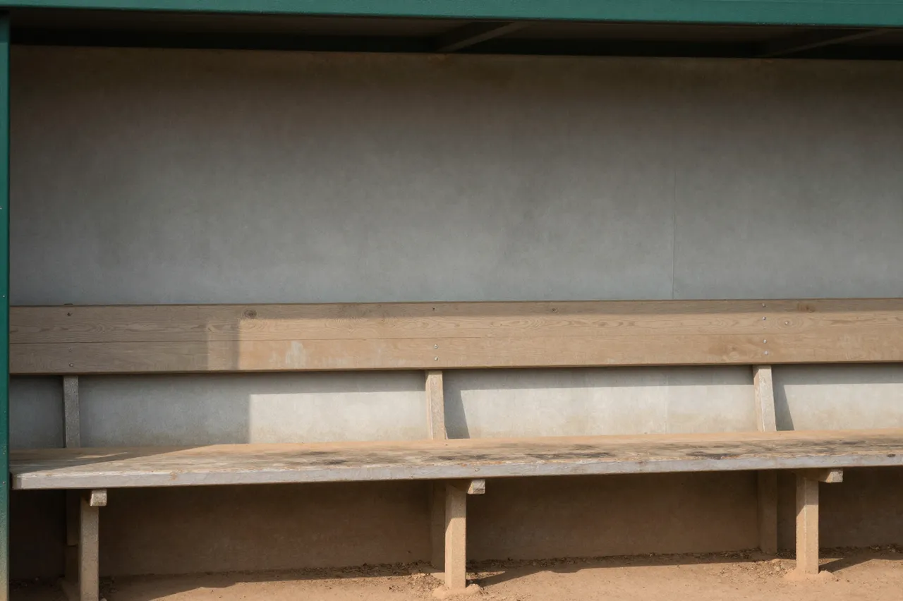 Unoccupied wooden and concrete dugout bench sits inside a small ballpark dugout in afternoon sunlight