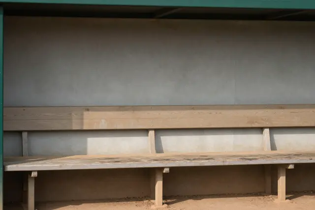 Unoccupied wooden and concrete dugout bench sits inside a small ballpark dugout in afternoon sunlight