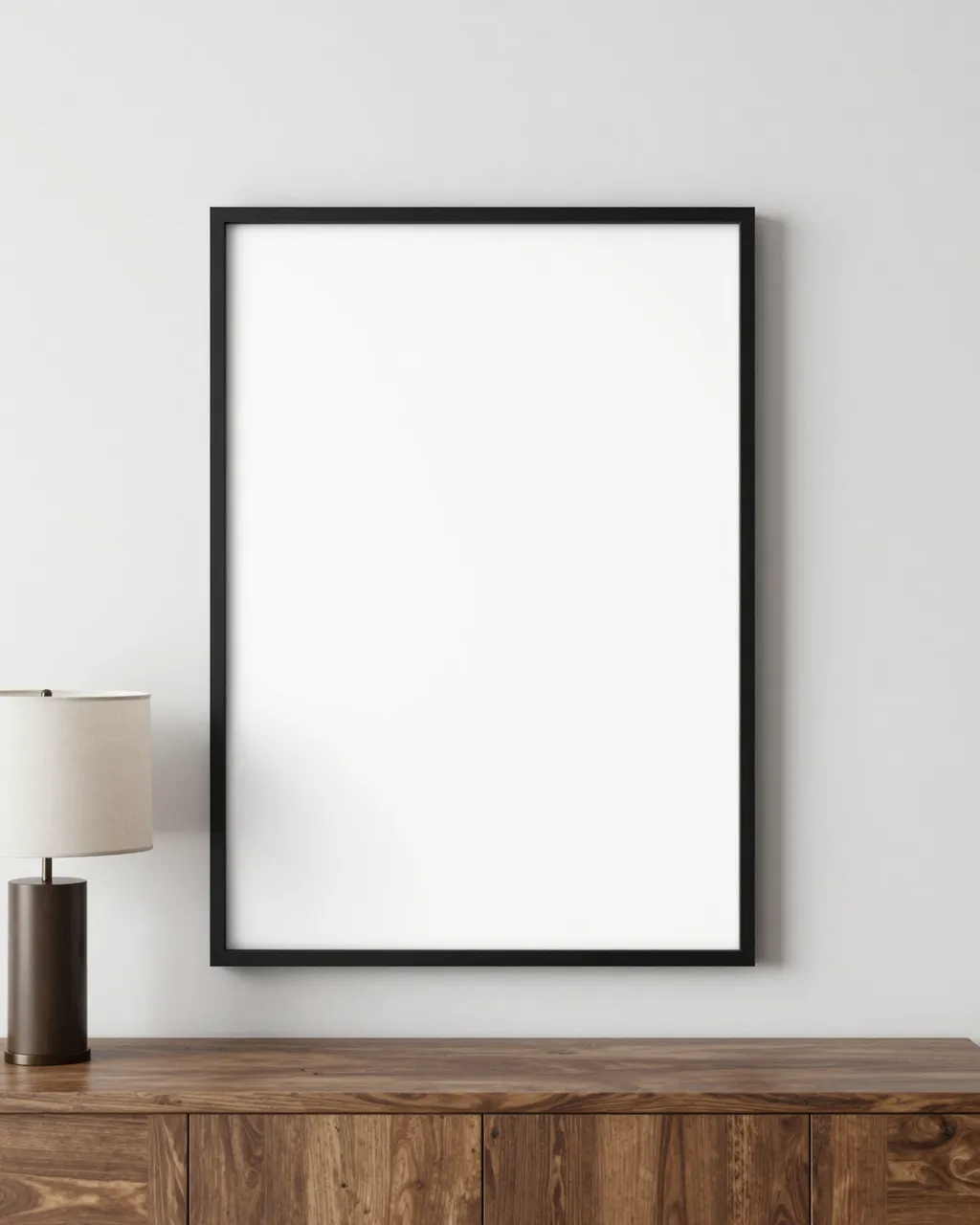 Blank thin black picture frame hangs above a walnut console table with a table lamp against a light white wall