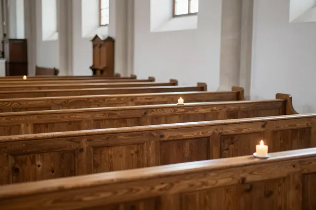 Rows of polished wooden church pews hold small lit candles beside light plaster walls and tall narrow windows.