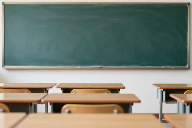 Rows of wooden school desks face a large blank green chalkboard in an empty classroom.