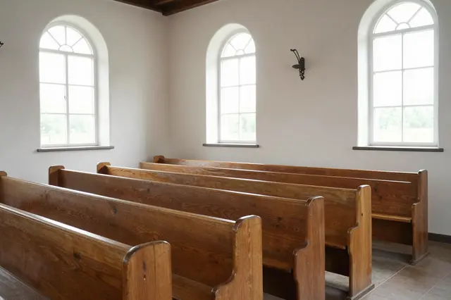 Rows of plain wooden pews fill a sunlit whitewashed church interior with arched windows.