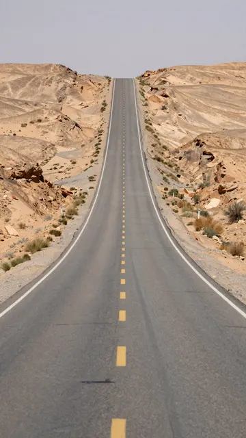 A straight empty paved desert road rises over rolling rocky hills under a pale midday sky