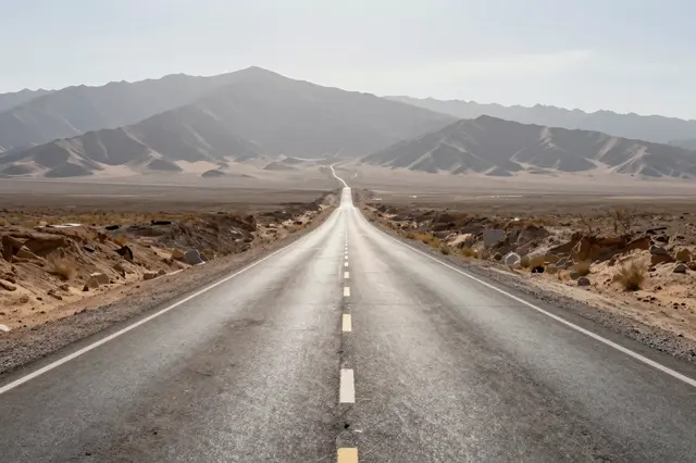 A straight empty paved desert road stretches toward hazy distant mountain ranges under a pale hot sky