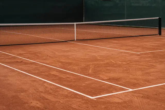 Swept reddish clay tennis court with net and white boundary lines, bordered by dark fence screening at dusk