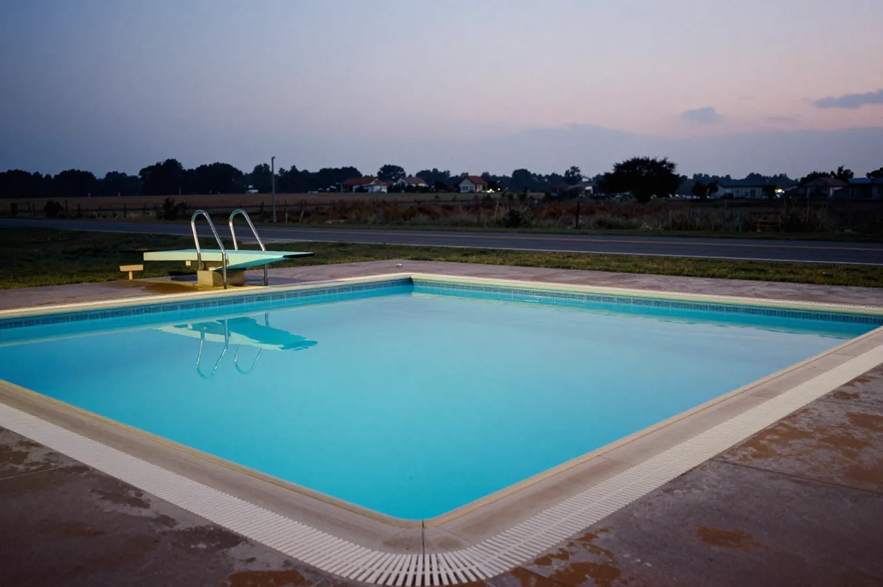 An empty in-ground outdoor pool with a diving board sits beside a rural road at soft dusk.