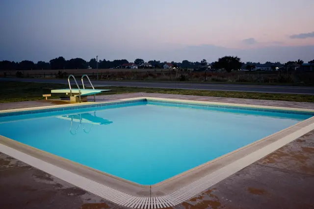 An empty in-ground outdoor pool with a diving board sits beside a rural road at soft dusk.