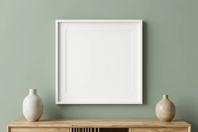 A blank white picture frame hangs above a wooden sideboard with two ceramic vases against a sage green wall.
