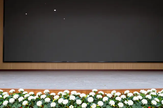 An empty stage decorated with rows of white flowers has falling confetti in front of a blank dark screen.