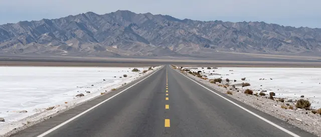 A straight empty paved highway stretches across a white salt flat toward distant rugged mountains