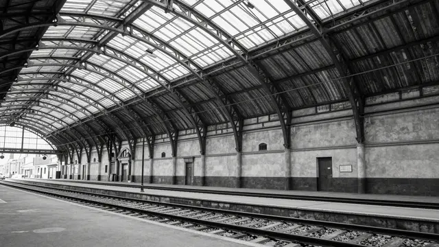 Black and white view of an empty old railway station with an arched glass and iron roof over tracks and platforms