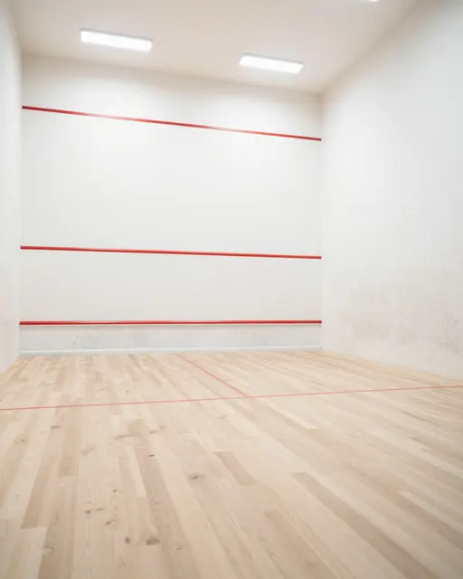 Unoccupied squash court with pale wooden flooring, white walls, and red court markings under ceiling lights