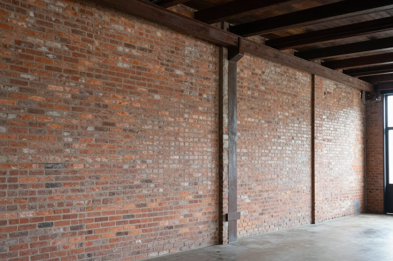 Unfurnished industrial loft with exposed red brick walls, dark wooden ceiling beams and a concrete floor