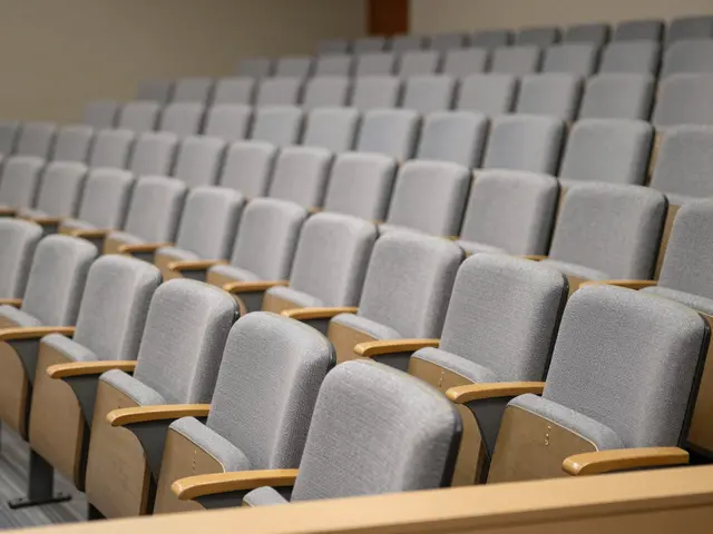 Steep tiered rows of empty gray upholstered lecture hall seats viewed from the back