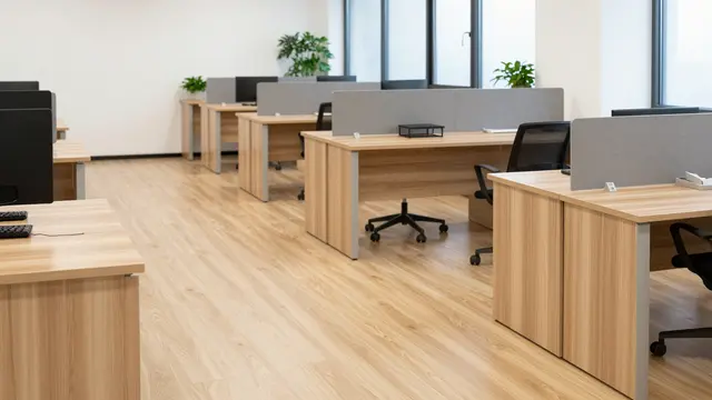Sunlit empty office lined with light wood desks, office chairs, and potted plants along large windows