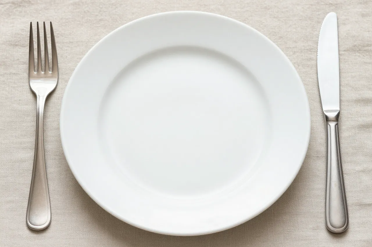 Top down view of an empty white dinner plate with a silver fork and knife placed on a beige linen tablecloth.