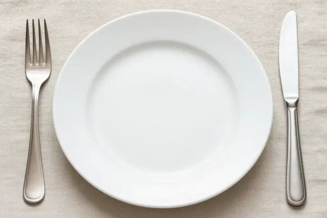 Top down view of an empty white dinner plate with a silver fork and knife placed on a beige linen tablecloth.