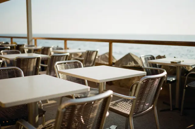 Unoccupied outdoor cafe tables and chairs overlook calm coastal water in warm afternoon light