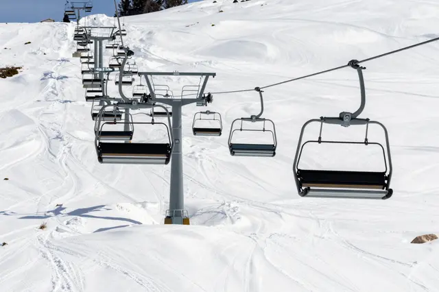 Empty ski lift chairs run up a groomed snowy mountain slope under clear sunny skies.