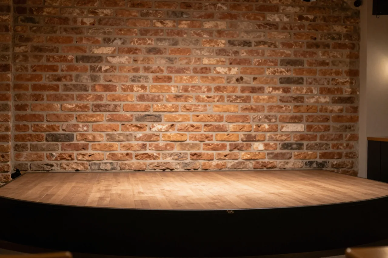Unoccupied wooden performance stage set against a warm exposed brick wall under soft interior lighting