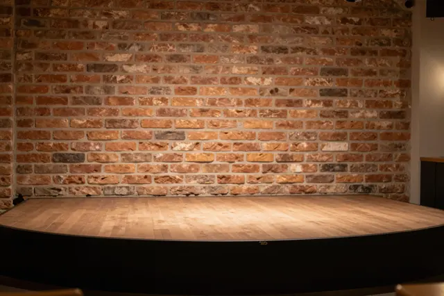 Unoccupied wooden performance stage set against a warm exposed brick wall under soft interior lighting