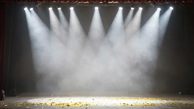 Bright white stage spotlights beam over an empty concert stage scattered with drifting gold confetti