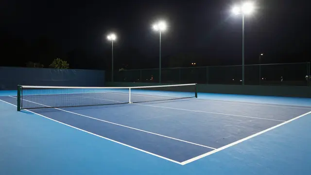 An empty blue hard tennis court sits illuminated by bright floodlights at night