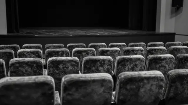 Black and white view of rows of velvet theater seats facing a dark empty stage