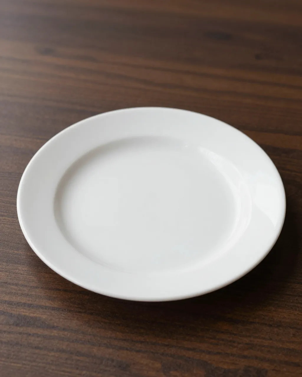 A plain empty white ceramic plate rests on a dark striped wood table with soft side lighting.
