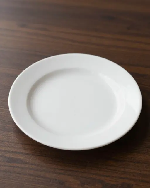 A plain empty white ceramic plate rests on a dark striped wood table with soft side lighting.