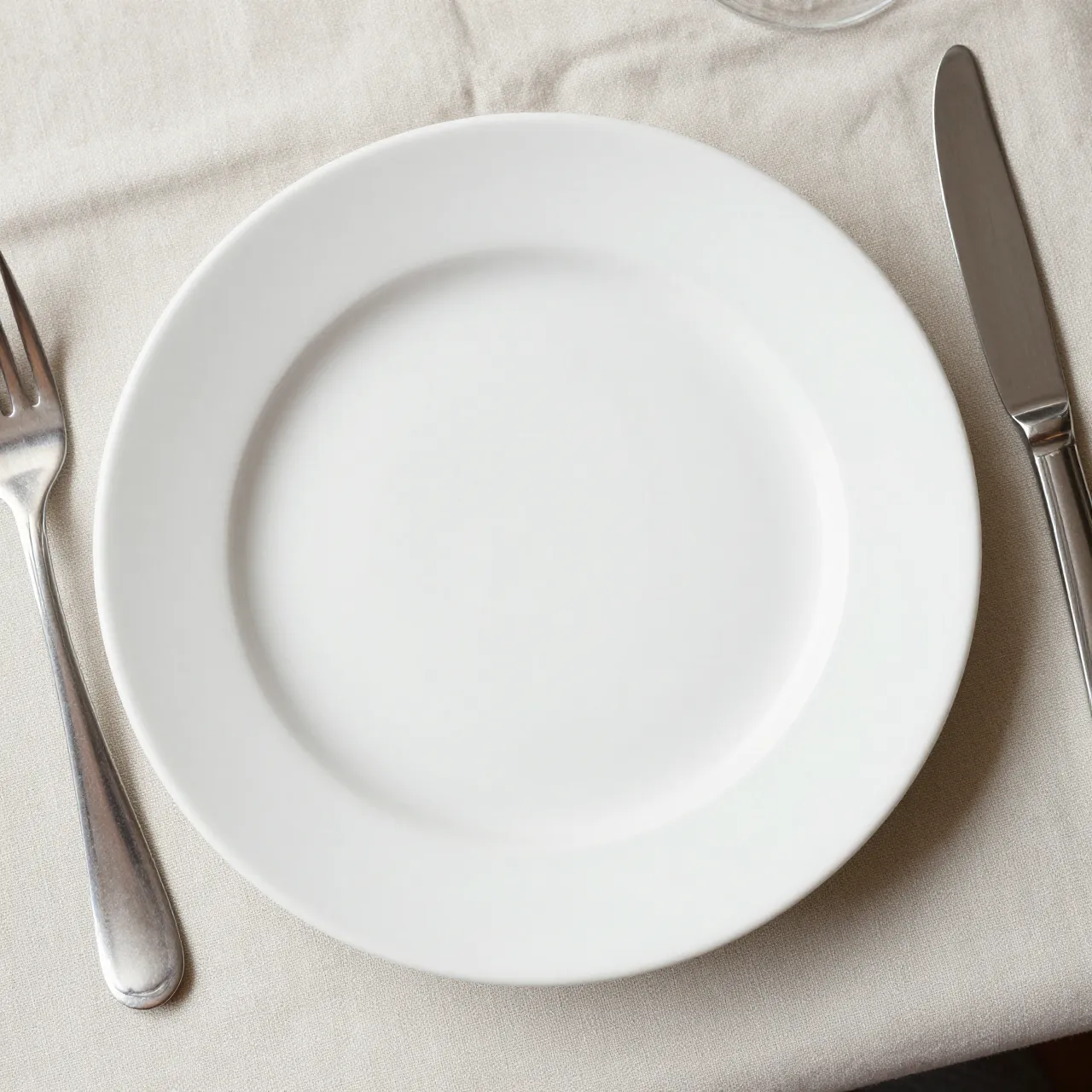 Top view of a blank white ceramic dinner plate with silver cutlery on a beige linen tablecloth.