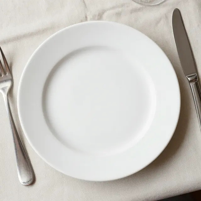 Top view of a blank white ceramic dinner plate with silver cutlery on a beige linen tablecloth.