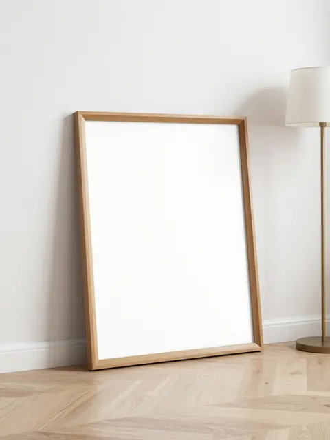 Blank wooden picture frame leans against a white wall next to a floor lamp on pale oak flooring