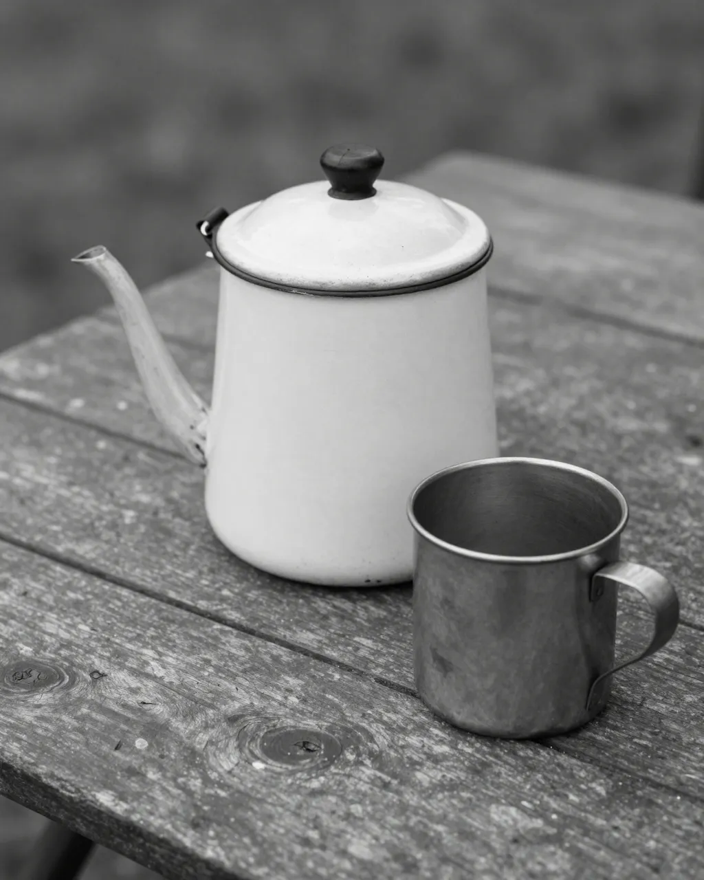 Black and white still life of an old enamel coffee pot and metal tin cup resting on a weathered wooden camp table