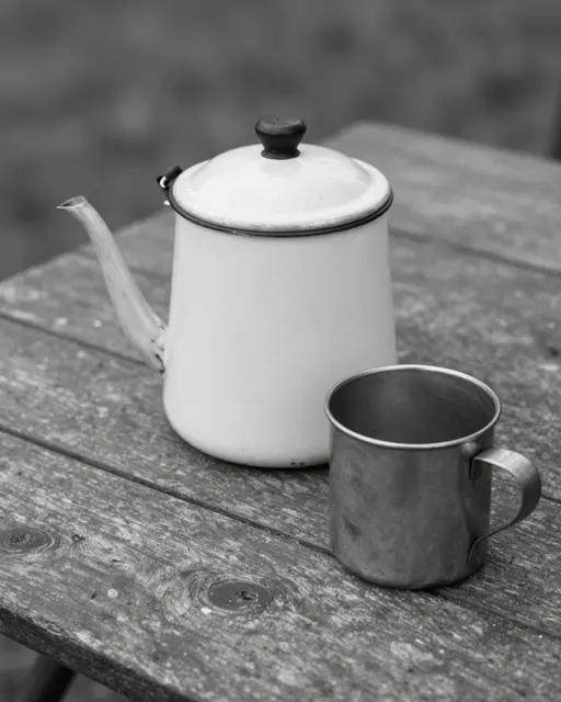 Black and white still life of an old enamel coffee pot and metal tin cup resting on a weathered wooden camp table