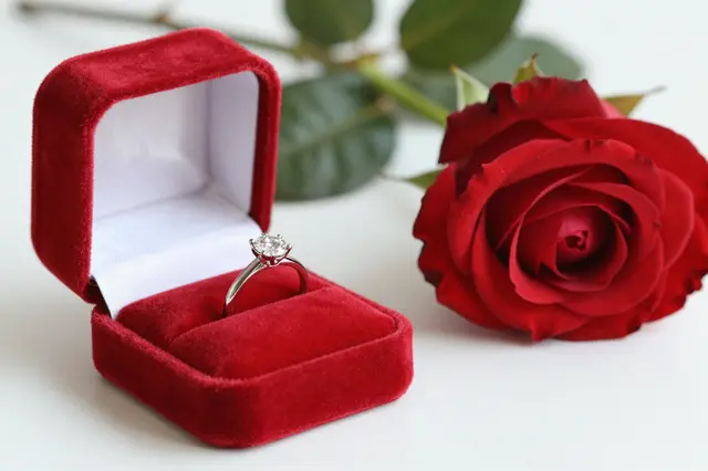 Open red velvet ring box holding a diamond engagement ring next to a fresh red rose on a white surface