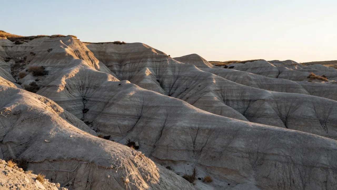 Warm sunrise light falls across layered eroded gray badlands hills under a pale sky