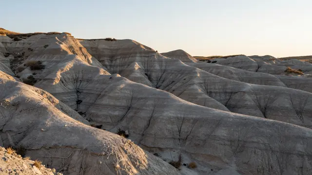 Warm sunrise light falls across layered eroded gray badlands hills under a pale sky
