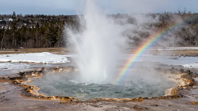 An erupting geyser sends spray upward, with a small rainbow visible in the mist over a snowy geothermal basin.