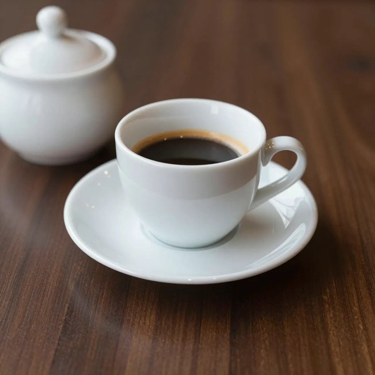 White espresso cup on a matching saucer sits beside a lidded sugar bowl on a dark walnut table.