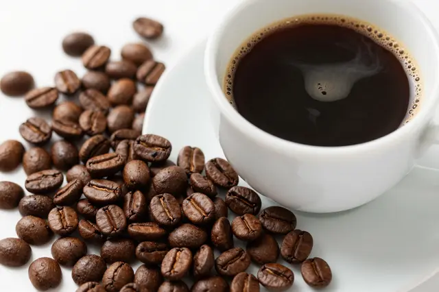 Close-up of a white ceramic espresso cup next to scattered roasted coffee beans on a light white surface.
