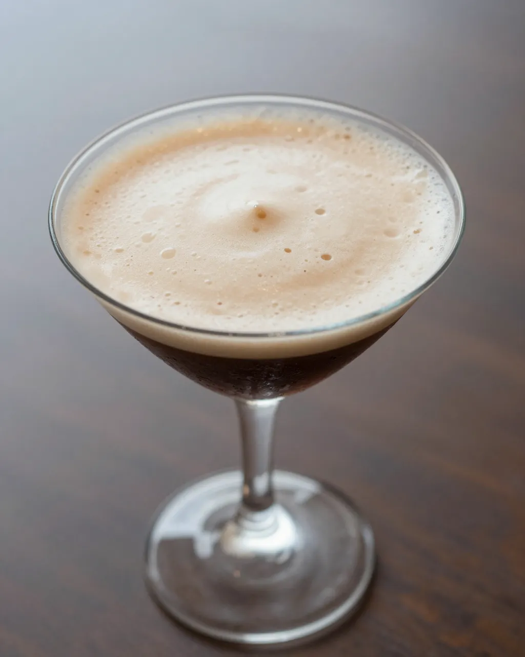 A frothy espresso martini cocktail served in a clear coupe glass resting on a dark wooden table.