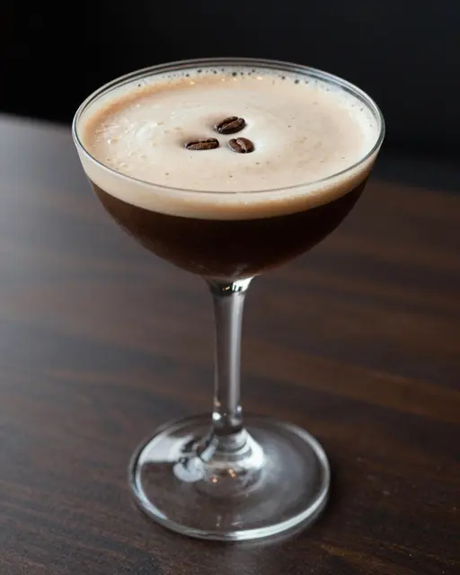 An espresso martini garnished with three coffee beans rests in a coupe glass on a dark wooden bar table.