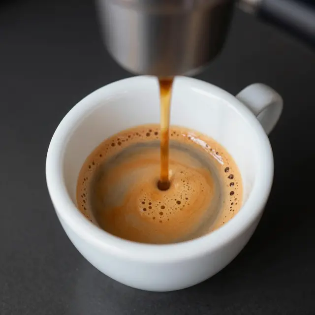 Fresh espresso pours from a portafilter into a small white cup topped with bubbly crema.