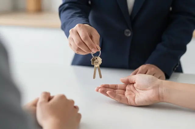 An estate agent extends house keys toward a waiting outstretched hand over a white table