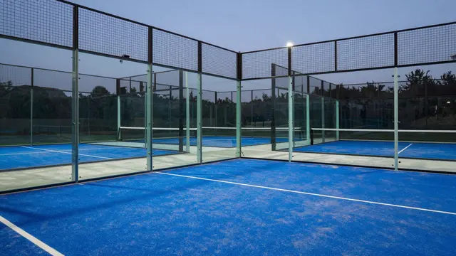 Empty padel court with glass walls and blue turf under soft evening twilight lighting