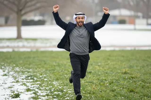 An excited Middle Eastern man runs toward the camera with raised fists on a snowy winter lawn.
