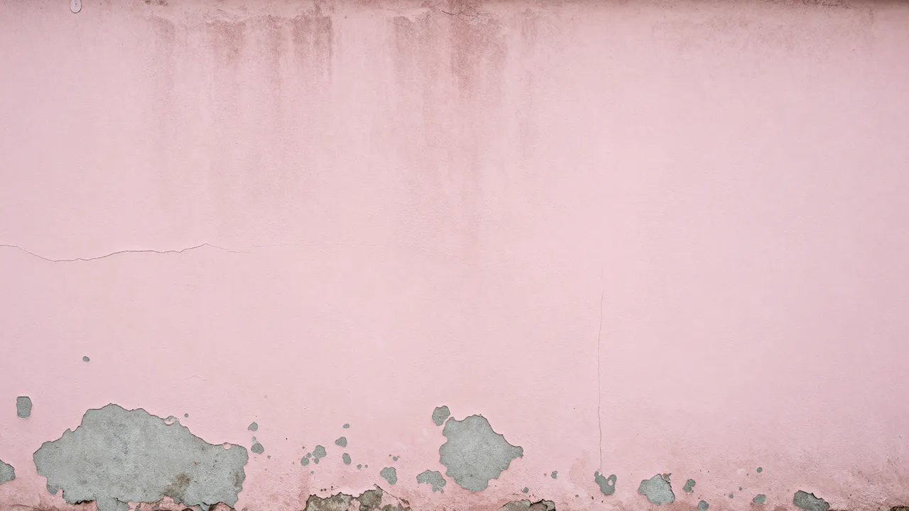 A weathered pink plaster wall with peeling paint revealing gray cement and fine cracks